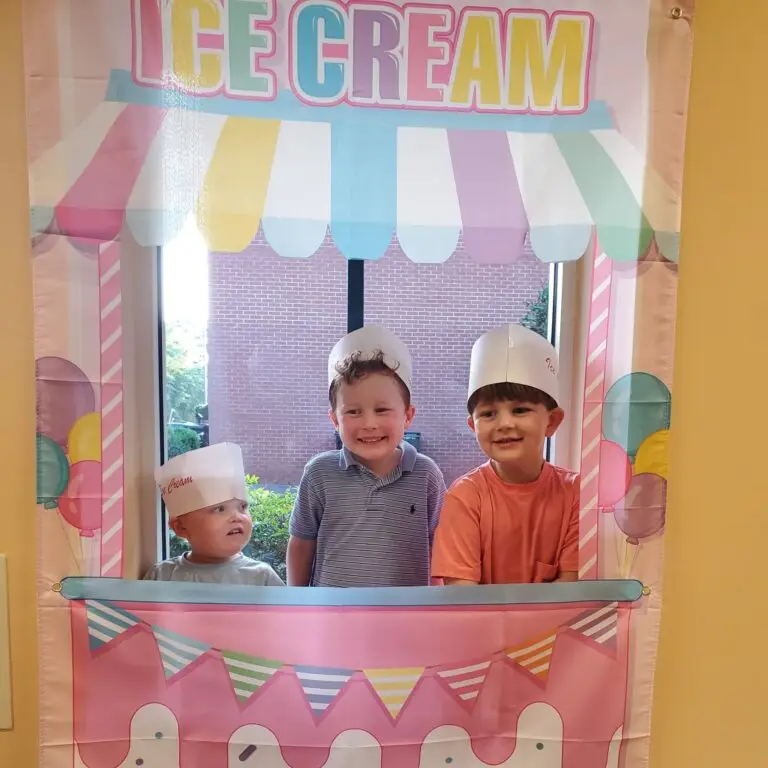 A toddler boy and two preschool boys pose with white hats on in temporary ice cream booth