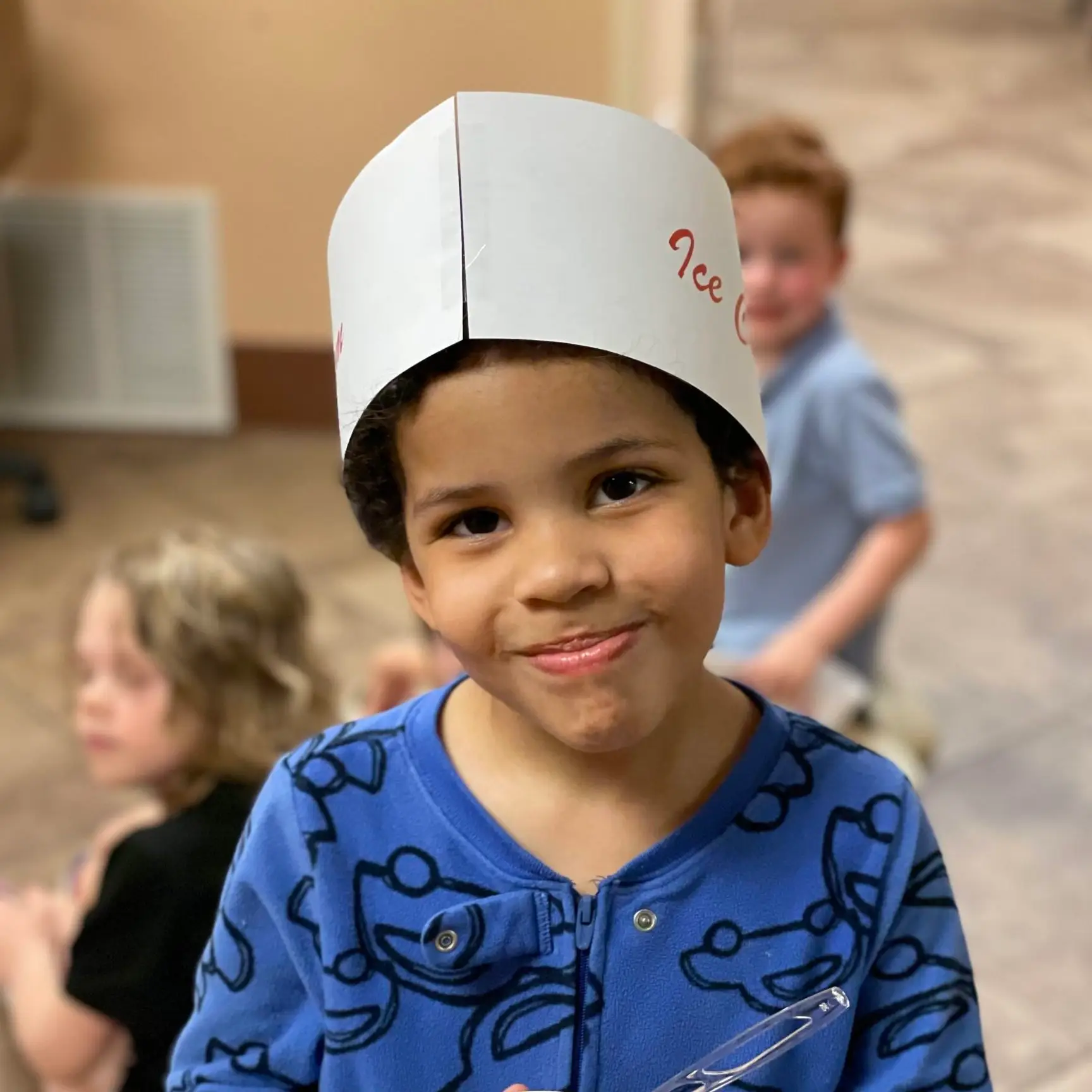 Little boy with dark curly hair under a white hat in blue pajamas holding a bowl of ice cream