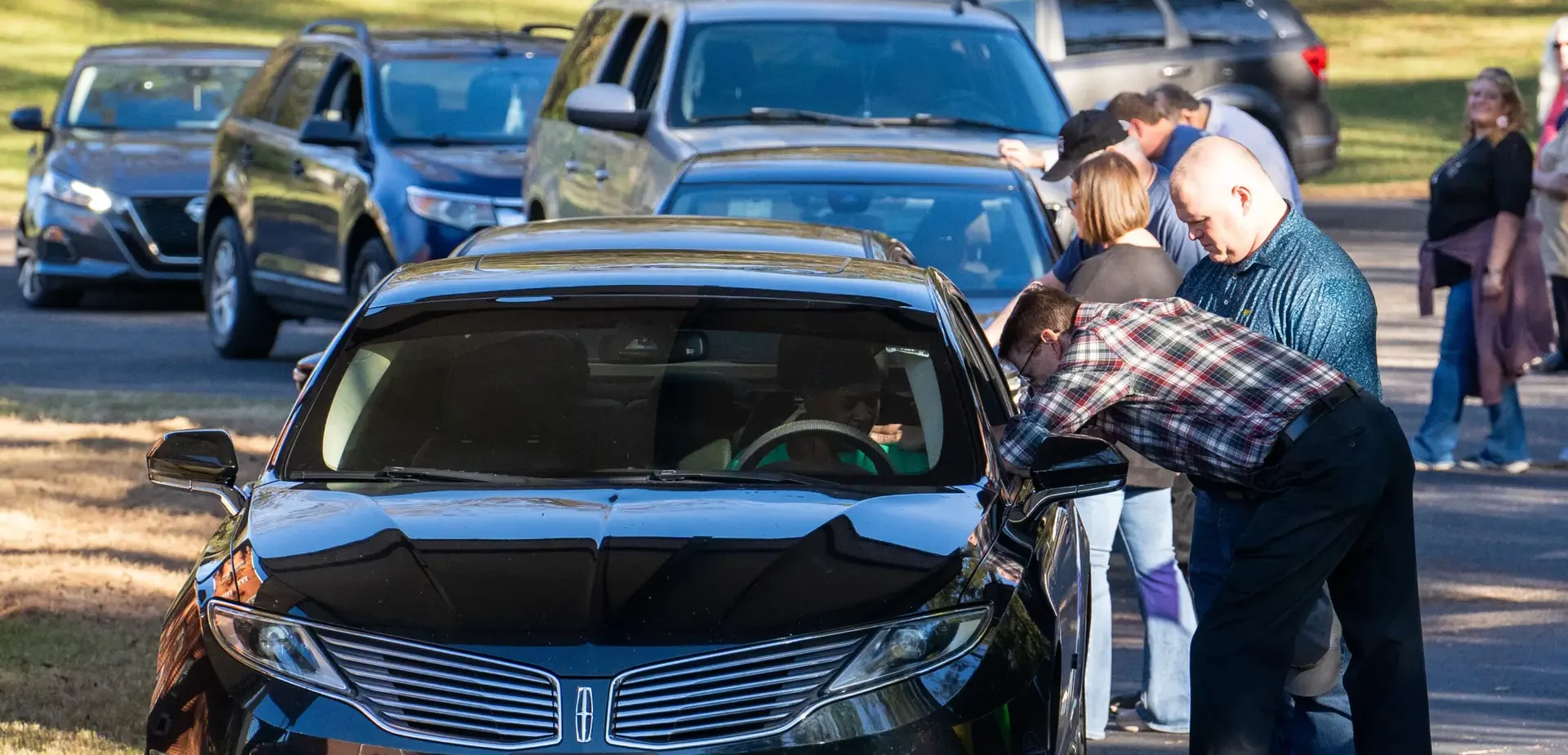 Community members sit in cars while church members stand beside their driver side window and pray for them at the Meal Kit Giveaway