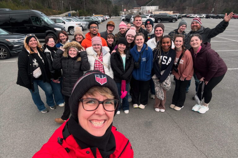 A woman in glasses is taking a selfie with a large group of students posing for a group photo behind her